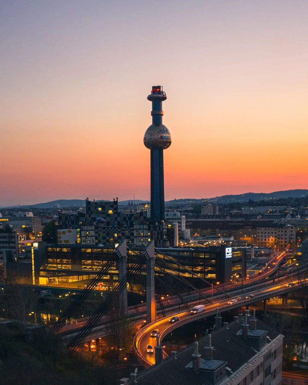 Stadtlandschaft bei Sonnenuntergang: Die kugelförmige Spitze der Müllverbrennungsannlage Spittelau glänzt vor dem Himmel, während die gewundenen Straßen darunter von Straßenlaternen beleuchtet werden.