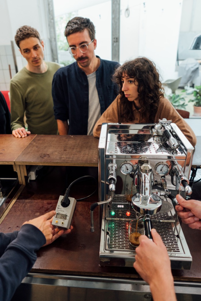 Menschen versammeln sich um eine Kaffeemaschine und beobachten jemanden, der mit einem Strommessgerät den Stromverbrauch misst.