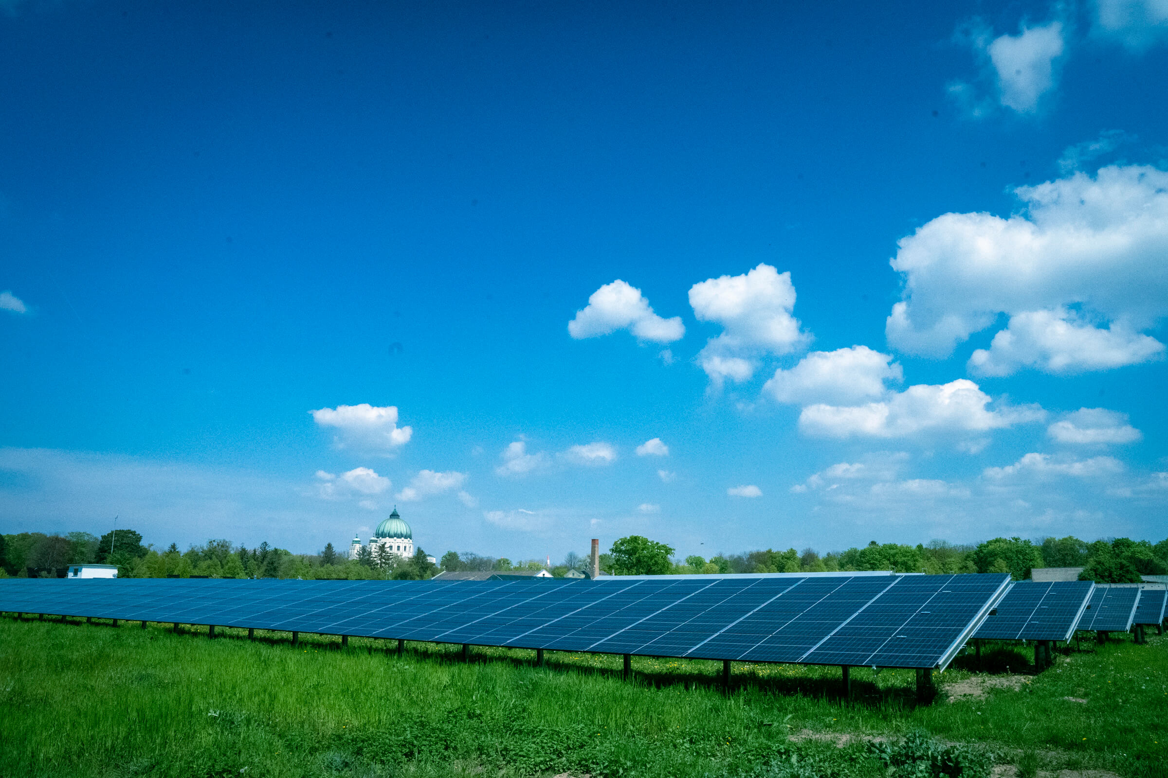Sonnenkollektoren auf einer grünen Wiese unter blauem Himmel mit vereinzelten Wolken. Im Hintergrund ist die Friedhofskirche des Zentralfriedhofs zu sehen.