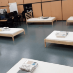 Zimmer mit mehreren Einzelbetten, jeweils mit gefalteter Bettwäsche darauf. Schreibtische und Stühle an den Wänden. Isolationsstationen in Spittelau, Flötzersteig und Simmeringer Haide.