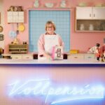 Eine Frau steht lächelnd in einer Retroküche und hält ein Buch in der Hand. Auf einem Neonschild steht „Vollpension“.