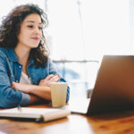 Frau mit lockigem Haar lächelt im Café ihren Laptop an, sitzt neben einem Notizbuch und einer Kaffeetasse auf einem Holztisch.