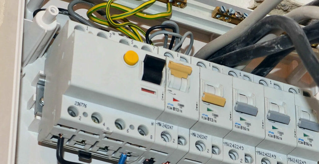 Nahaufnahme einer elektrischen Leistungsschaltertafel mit mehreren Schaltern und beschrifteten Kabeln. Links befindet sich der Testknopf.