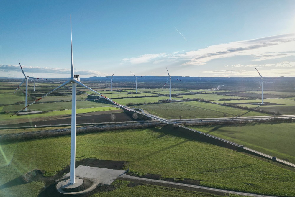 Windräder des Windparks Trumau an einem sonnigen Tag in einer flachen, grünen Landschaft mit fernen Bergen im Hintergrund.