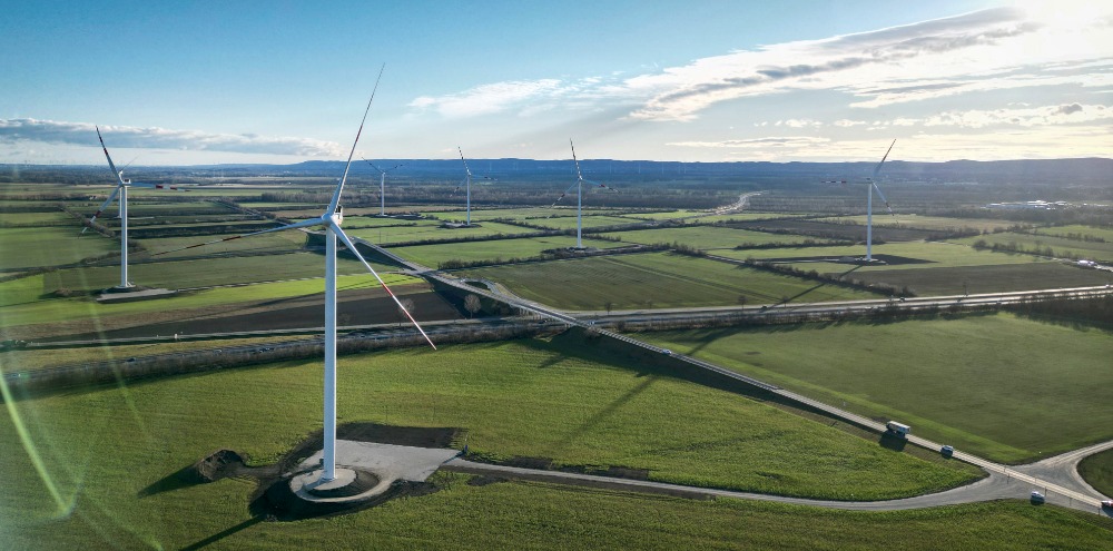 Windräder des Windparks Trumau an einem sonnigen Tag in einer flachen, grünen Landschaft mit fernen Bergen im Hintergrund.