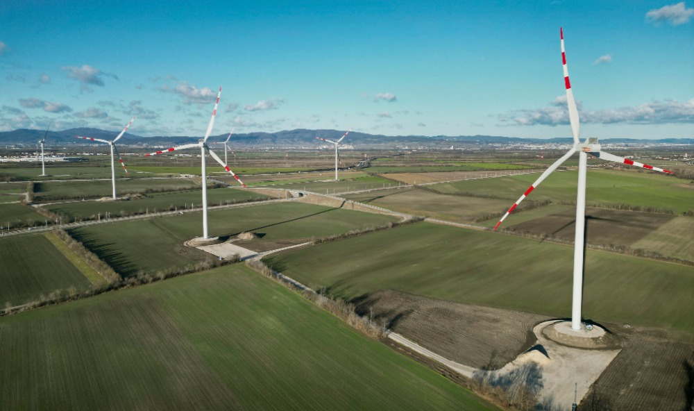 Windräder des Windparks Trumau an einem sonnigen Tag in einer flachen, grünen Landschaft mit fernen Bergen im Hintergrund.