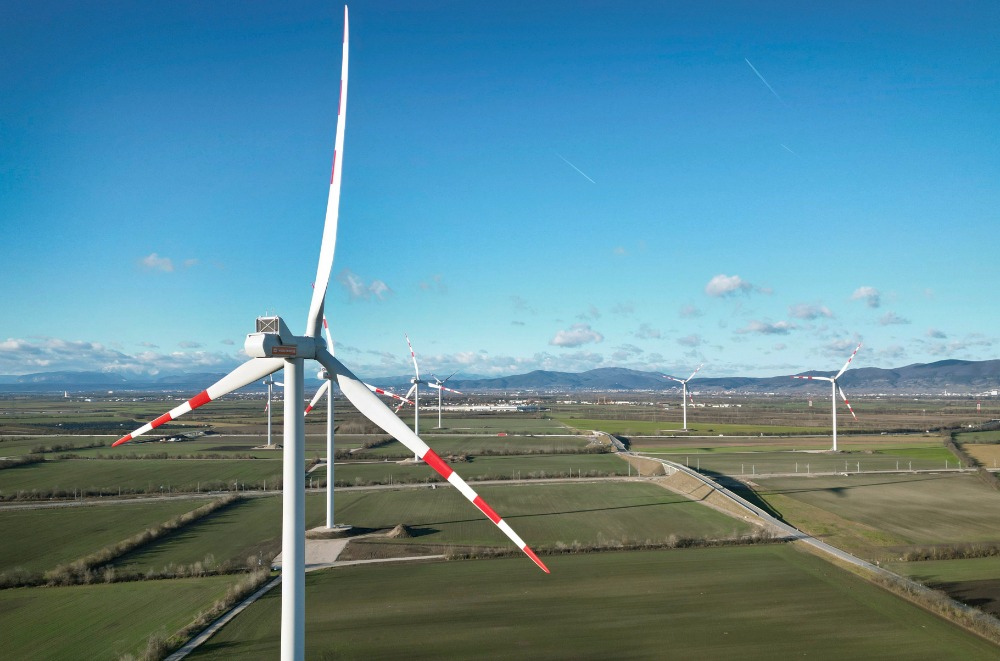 Windturbinen in einer weitläufigen grünen Landschaft unter einem klaren blauen Himmel mit Bergen in der Ferne.