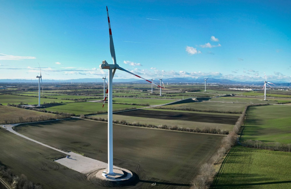 Windräder des Windparks Trumau an einem sonnigen Tag in einer flachen, grünen Landschaft mit fernen Bergen im Hintergrund.