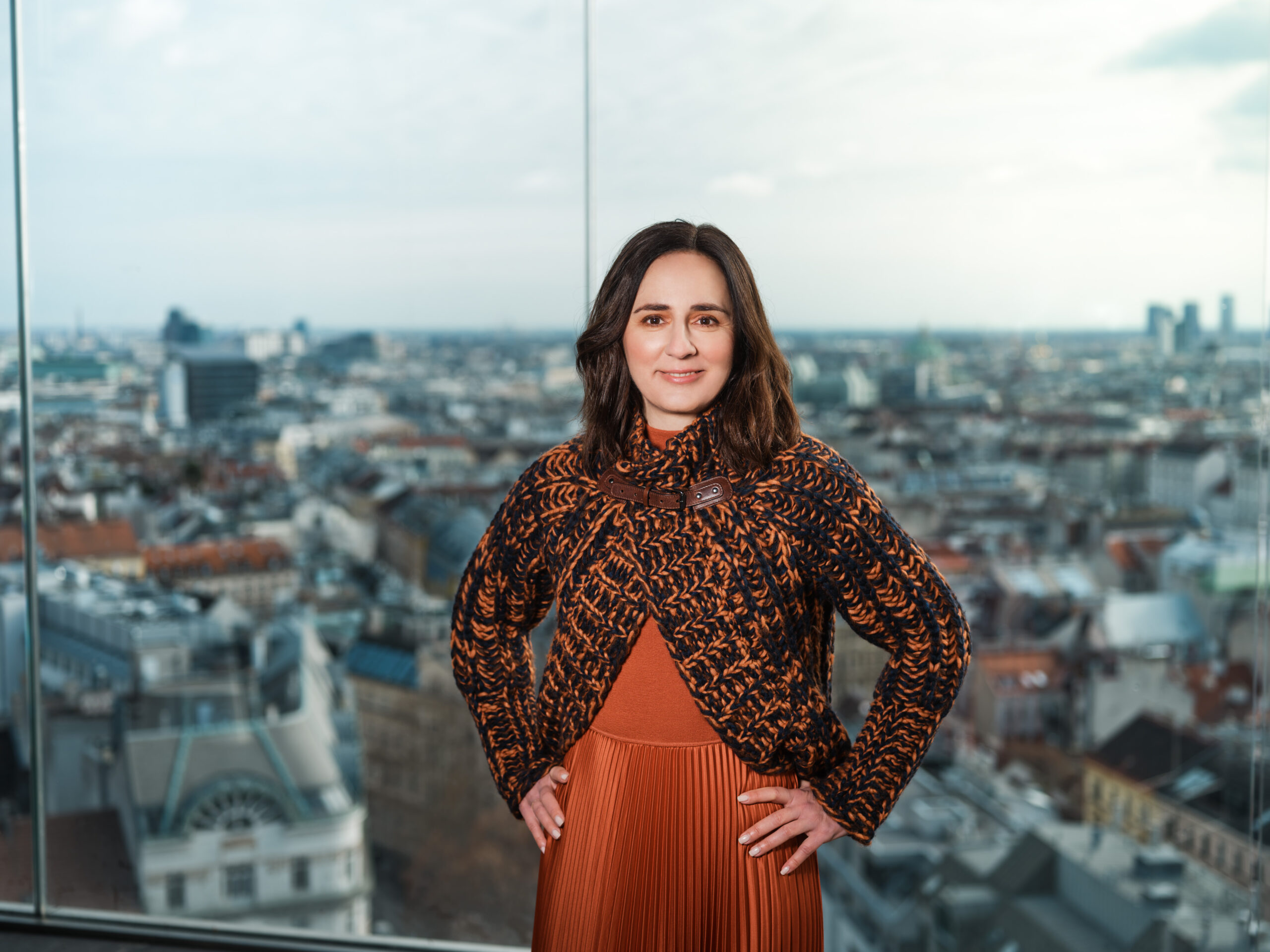 Alma Kahler steht selbstbewusst in einem gemusterten Pullover da, während sich durch das Glasfenster hinter ihr die Wiener Stadtlandschaft entfaltet.