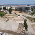 Auf der Baustelle im Village im Dritten herrscht geschäftiges Treiben mit Maschinen und Kieshaufen, vor der Kulisse malerischer Wohnhäuser und üppiger Bäume.