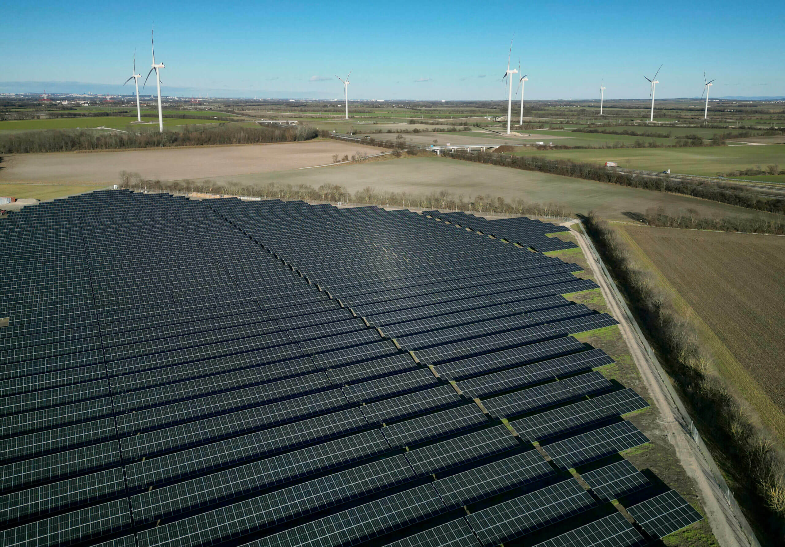 Luftaufnahme einer großen Photovoltaikanlage des Hybridkraftwerks Trumau mit Windturbinen im Hintergrund über einer ländlichen Landschaft.