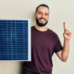 Lächelnder Mann hält ein Solarpanel und zeigt mit seinem Finger nach oben vor einem einfarbigen Hintergrund.