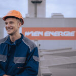 Jakob Königshofer, ein Wien Energie Mitarbeiter in blauer Uniform und Schutzhelm, steht stolz vor einem Gebäude, das das Schild „WIEN ENERGIE“ schmückt.