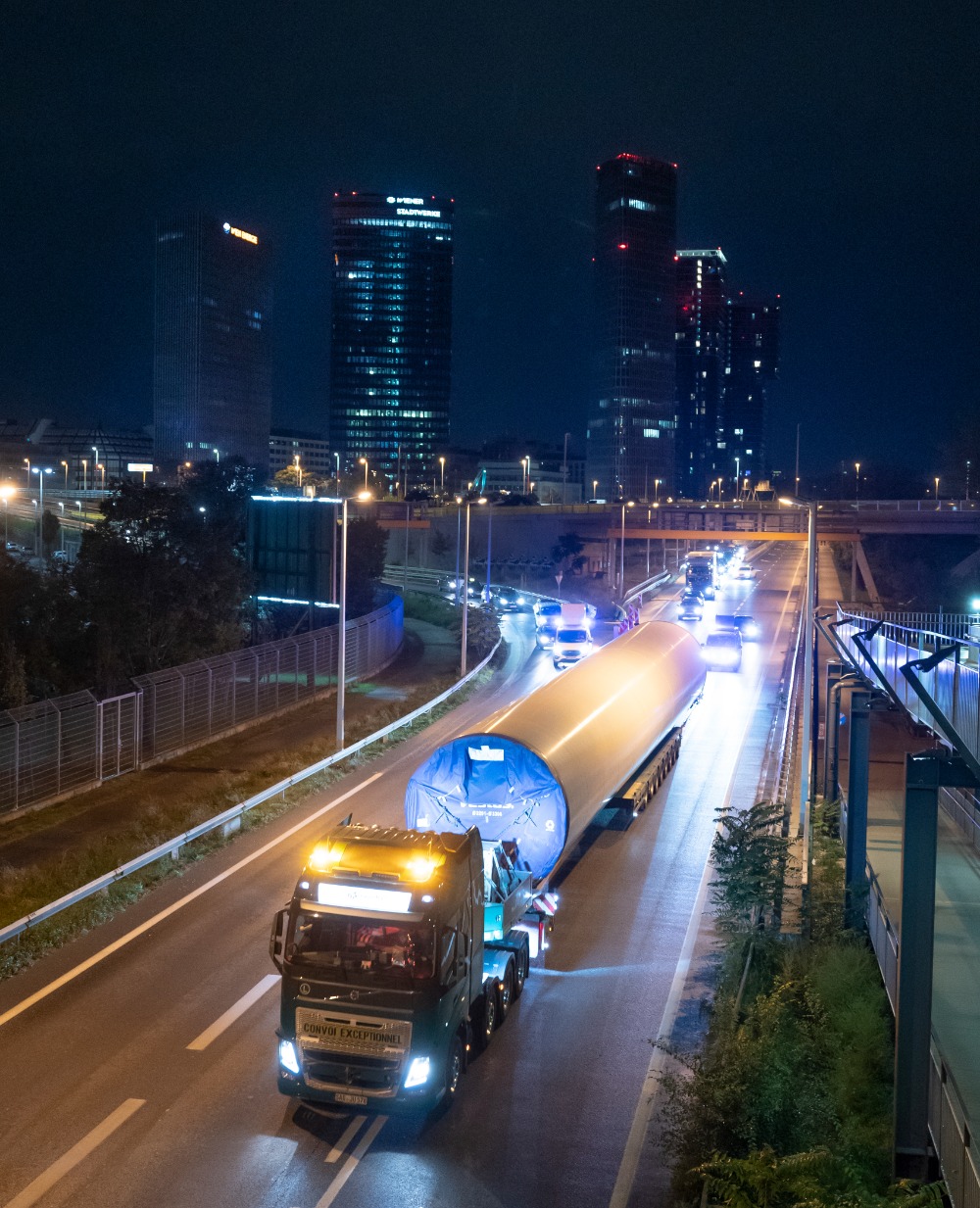 Ein Lastwagen transportiert ein großes zylindrisches Objekt für ein Windrad auf einer Autobahn bei Nacht, im Hintergrund sind Wolkenkratzer zu sehen.