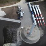 Luftaufnahme einer Windkraftanlagen-Baustelle mit Turbinenblättern auf dem Boden und einem zentralen Turmsockel.