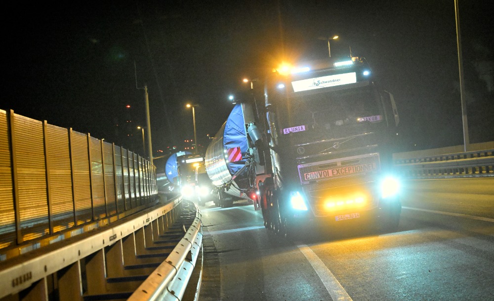 Ein LKW transportiert nachts große Rotorblätter einer Windkraftanlage auf einer Autobahn, beleuchtet von Scheinwerfern und Straßenlaternen.