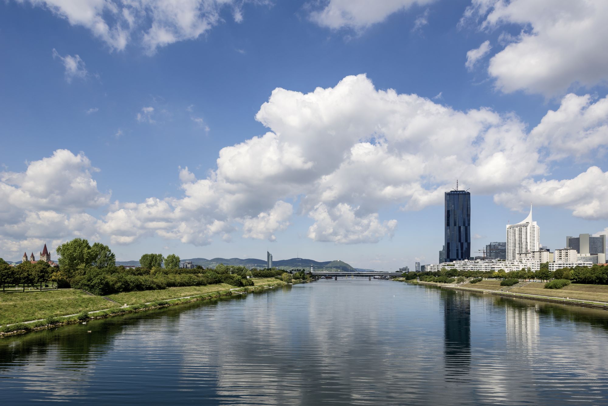 Donau mit Wiener Skyline, modernen Wolkenkratzern und Brücke unter blauem Himmel mit Wolken. Grasbewachsene Ufer säumen den Fluss.
