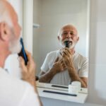 Ein älterer Mann mit grauem Haar rasiert sich vor einem Badezimmerspiegel und hält einen Rasierer an seinen Bart.