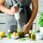 Person mixt in einer Küche grüne Soße. Auf dem Tisch liegen Kräuter, Zitrone und Käse.