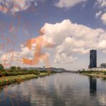 Die Skyline in Wien mit der ruhigen Donauinsel im Vordergrund und einem geometrischen orangefarbenen Muster (Business Run Logo) auf der linken Seite.