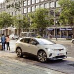 Elektroauto auf einer Stadtstraße geparkt, Menschen mit Gepäck in der Nähe, Gebäude und Bäume im Hintergrund.