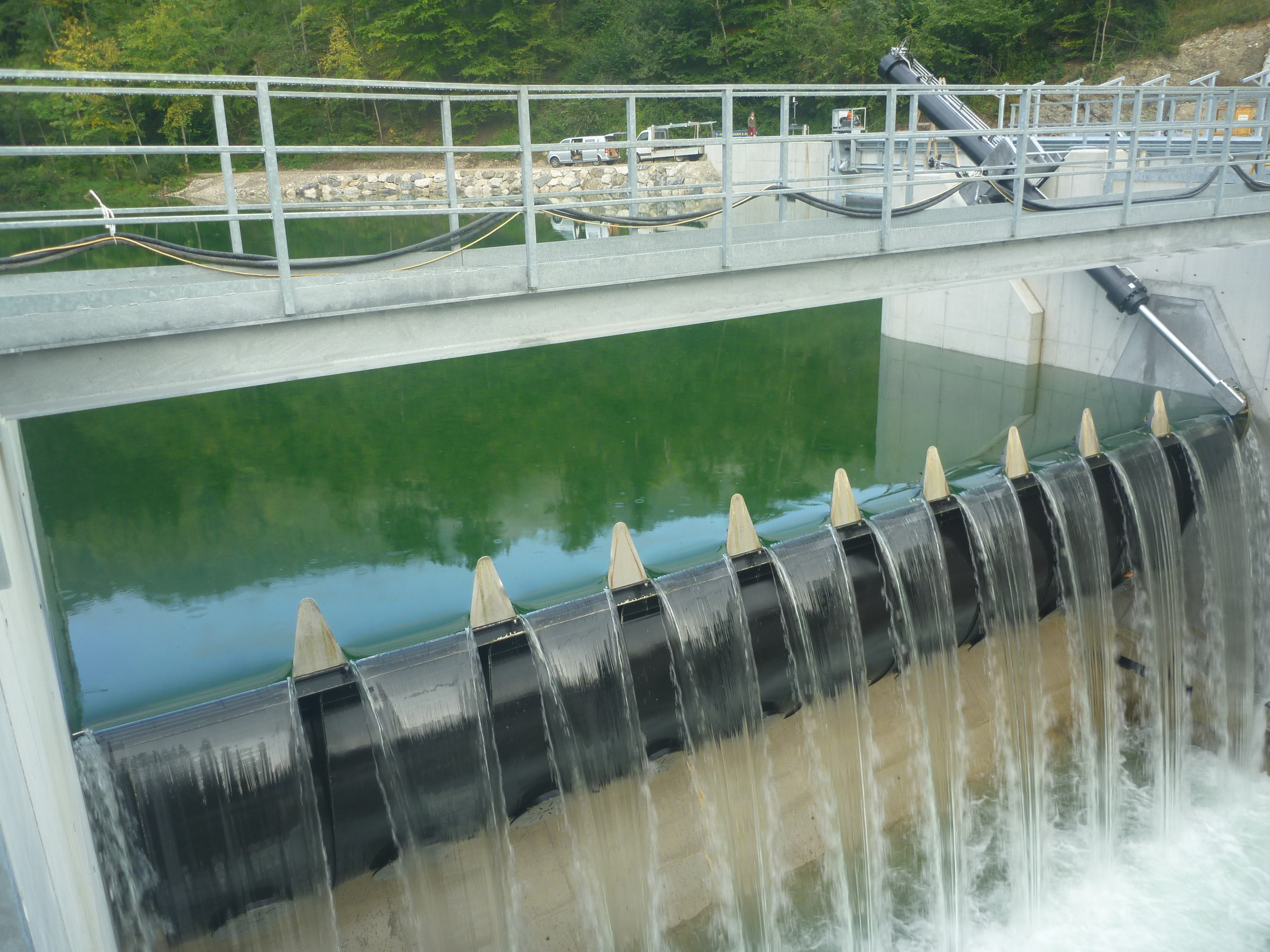 Ein kleiner Damm, über dessen aufblasbare Gummibarrieren Wasser fließt, während sich hinter dem Bauwerk grünes Wasser sammelt – eine Konstruktion, die an das innovative Design des Kraftwerks Opponitz erinnert.