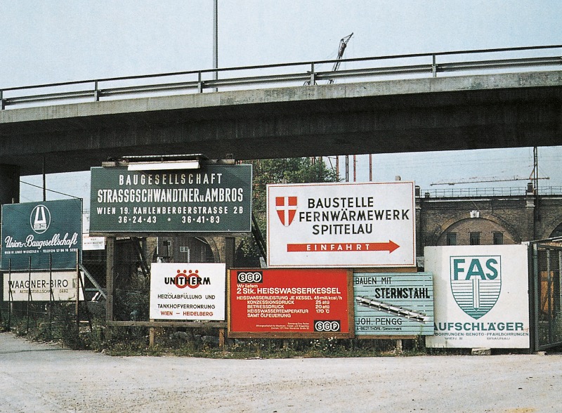 Unter der Brücke herrscht auf einer Baustelle reges Treiben. Sie ist mit Firmenschildern und Logos geschmückt, darunter prominent die „Baustelle Fernwärmewerk Spittelau“.
