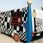 Draußen parkt ein farbenfroher Fernwärme-Bus mit Schachbrettmuster, kräftigem Textdesign und auffälligen roten Akzenten.