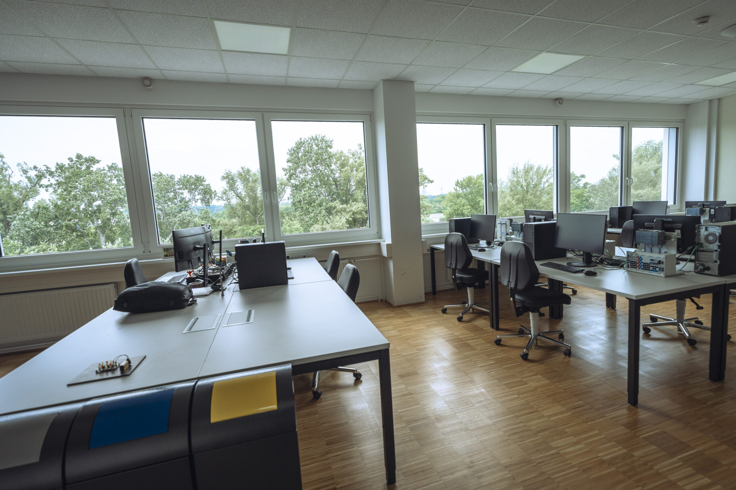 Leeres Schulungszimmer mit Schreibtischen, Computern und Stühlen neben großen Fenstern mit Blick auf Bäume.