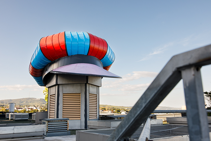 Eine farbenfrohe, abstrakte Dachkonstruktion, die vor einem klaren blauen Himmel an das "Hundertwasser-Kapperl" erinnert.