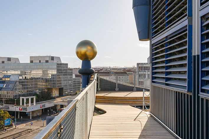 Blick vom Dach mit Gehweg, goldener Kugel und umgebender Stadtlandschaft unter klarem Himmel. Erlebnispfad der Spittelau.