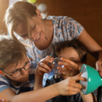 Eine Frau und zwei Kinder montieren lächelnd und konzentriert in einem warm beleuchteten Raum eine Lampe.