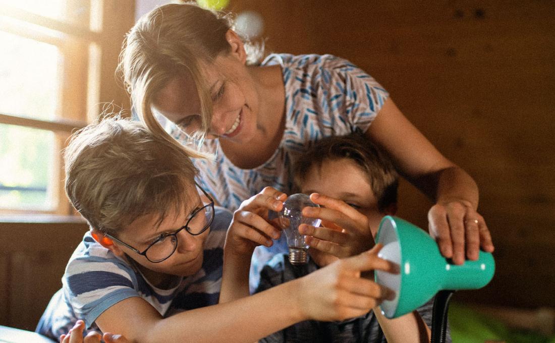 Eine Frau und zwei Kinder montieren lächelnd und konzentriert in einem warm beleuchteten Raum eine Lampe.