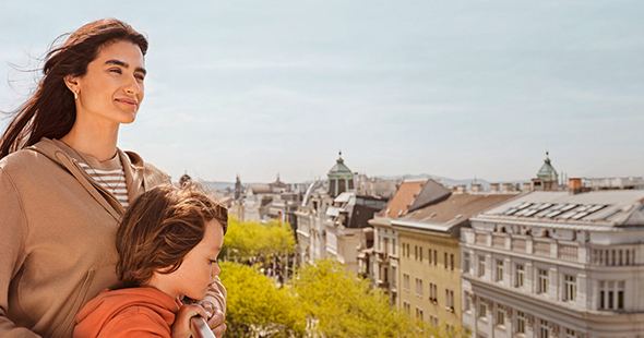 Frau und Kind betrachten die Wiener Stadtlandschaft mit historischen Gebäuden und Bäumen unter einem klaren Himmel.