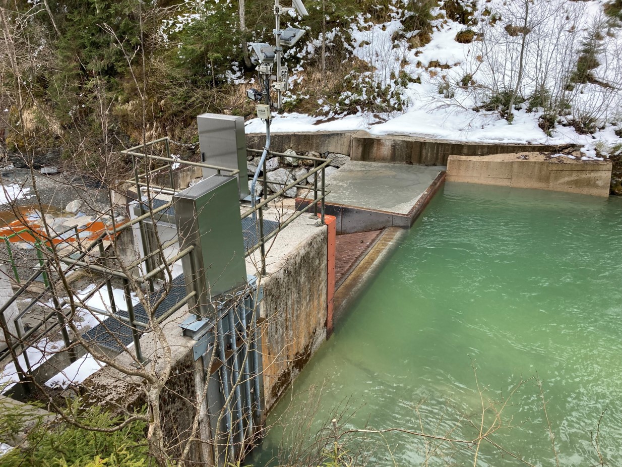 Wasserkraftwerk Mühlbach mit türkisfarbenem Wasser, umgeben von Schnee und Bäumen, mit Geräten und Betonstrukturen.