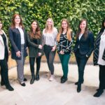 Sieben Wien Energie Frauen, die den Weltfrauentag feiern, stehen stolz vor einer leuchtend grünen Pflanzenwand und lächeln alle herzlich in ihrer lässig-eleganten Kleidung.