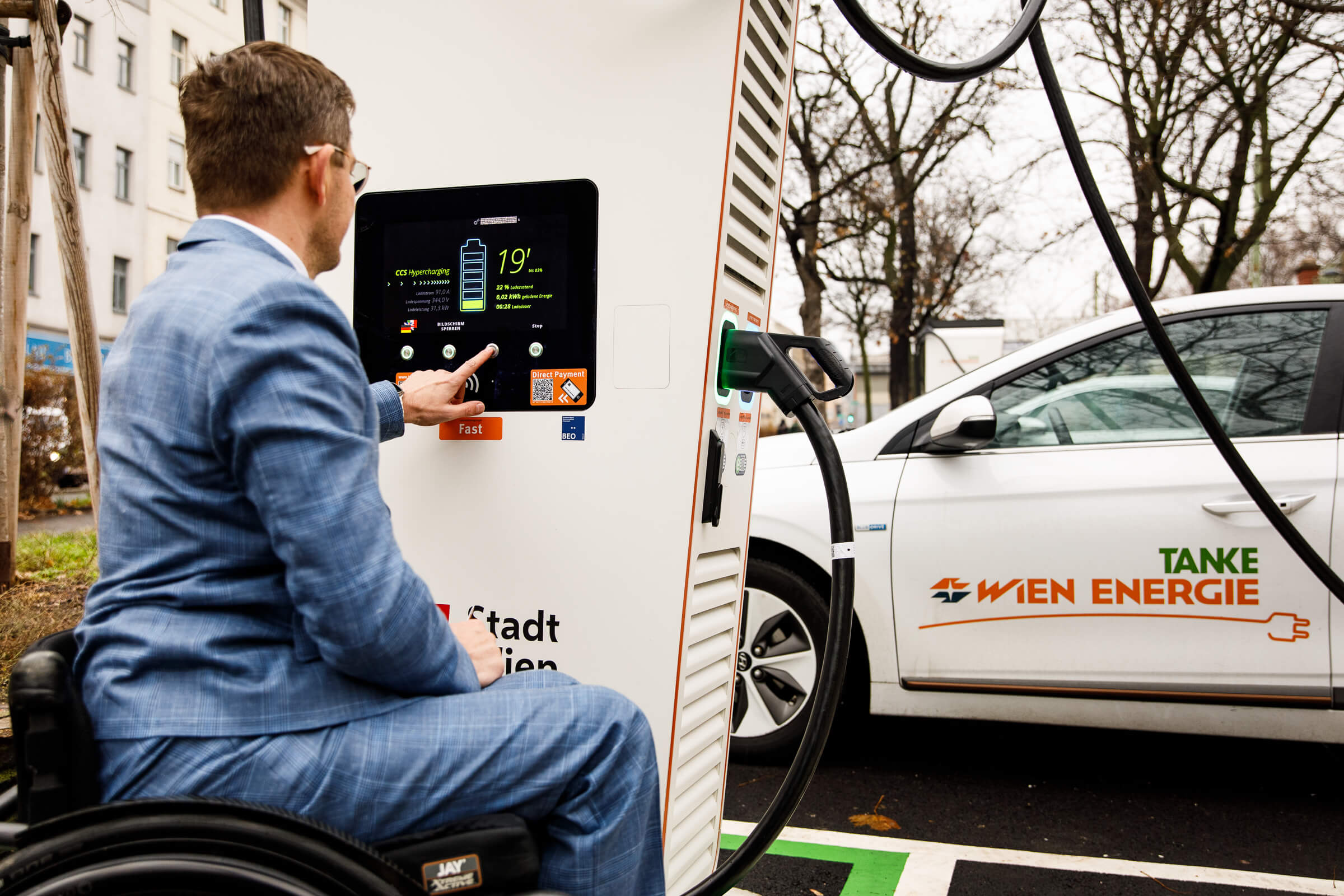 Ein Mann im Rollstuhl bedient neben einem Auto von Wien Energie eine Schnellladestation für Elektrofahrzeuge.