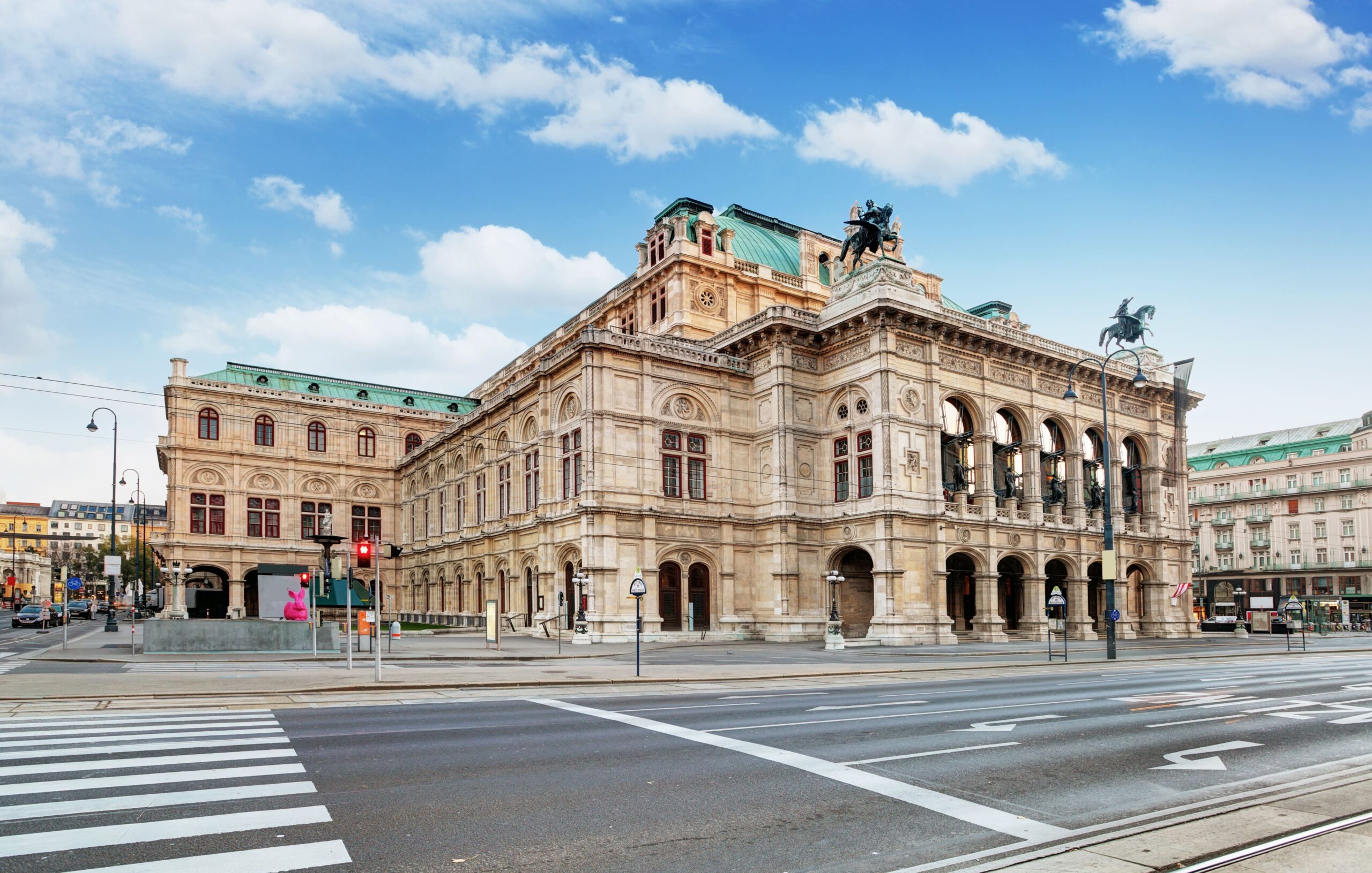 Die historische Wiener Staatsoper steht majestätisch in einer Stadtstraße und ihre kunstvolle Architektur glänzt unter einem blauen, wolkenverhangenen Himmel.
