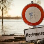 Ein „Hochwasser“-Schild vor einer überfluteten Landschaft mit kahlen Bäumen und einer unter Wasser liegenden Straße.
