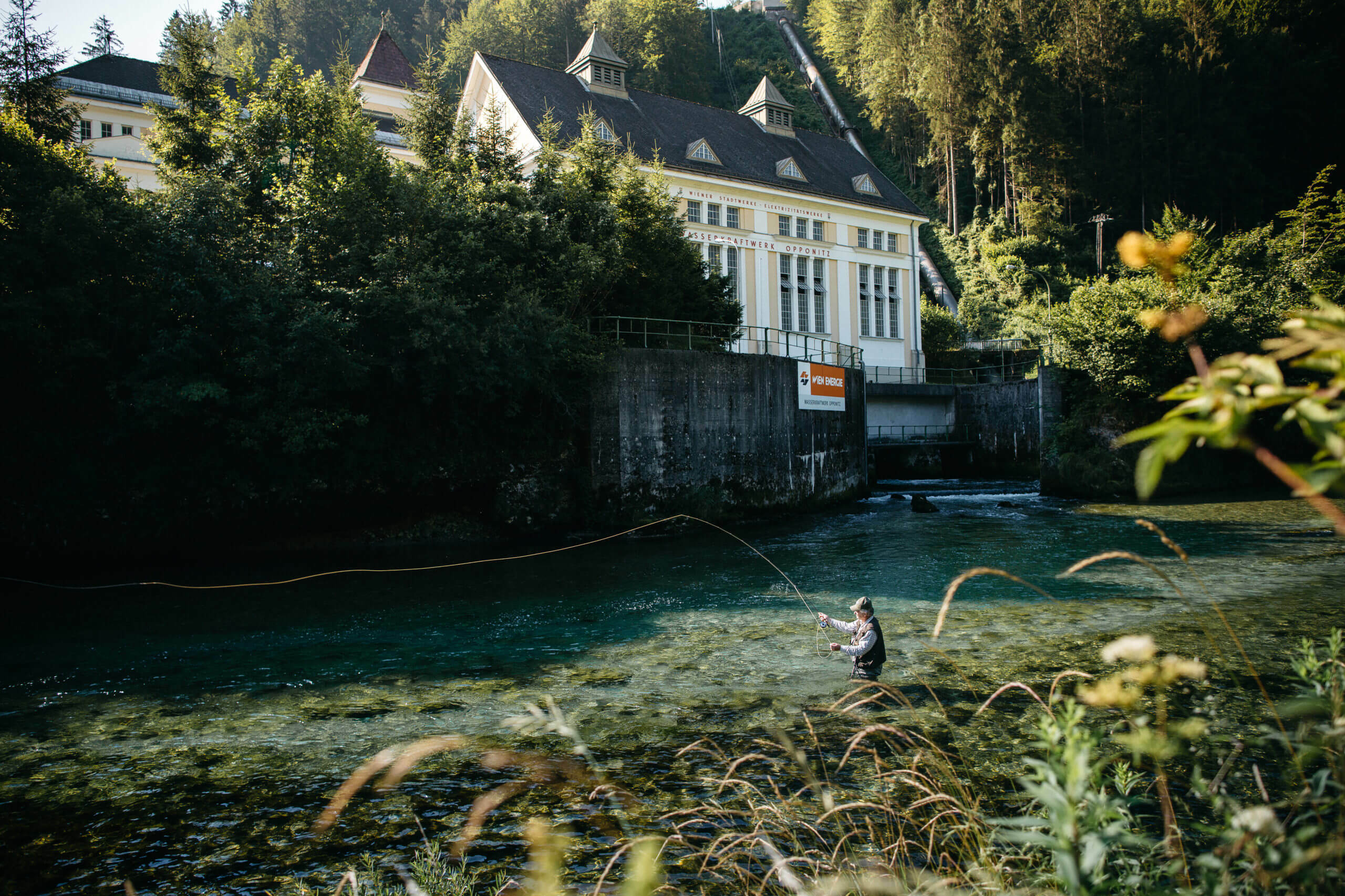 Angler beim Fliegenfischen in einem klaren Fluss in der Nähe des Wasserkraftwerkes Opponitz, umgeben von üppigem Grün und Bäumen.