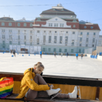 Eine Person in einem gelben Mantel sitzt auf einer Bank und bindet Schlittschuhe fest. Im Hintergrund sind eine Eisbahn und ein großes Gebäude, das Rathaus, zu sehen.