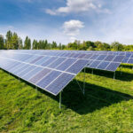 Ein Feld aus Photovoltaikmodulen in Traiskirchen glitzert unter einem teilweise bewölkten Himmel, im Hintergrund ragen hohe Bäume auf.