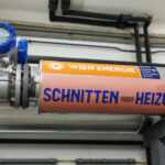 Ein Metallrohr der Fernwärme-Übergabestation mit der Aufschrift "SCHNITTEN HEIZUNG" und dem Wien Energie-Logo sowie einem angebrachten Messgerät.