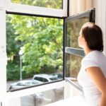 Eine Person im weißen Hemd blickt durch ein offenes Fenster nach draußen und sieht dabei Grün und geparkte Autos.