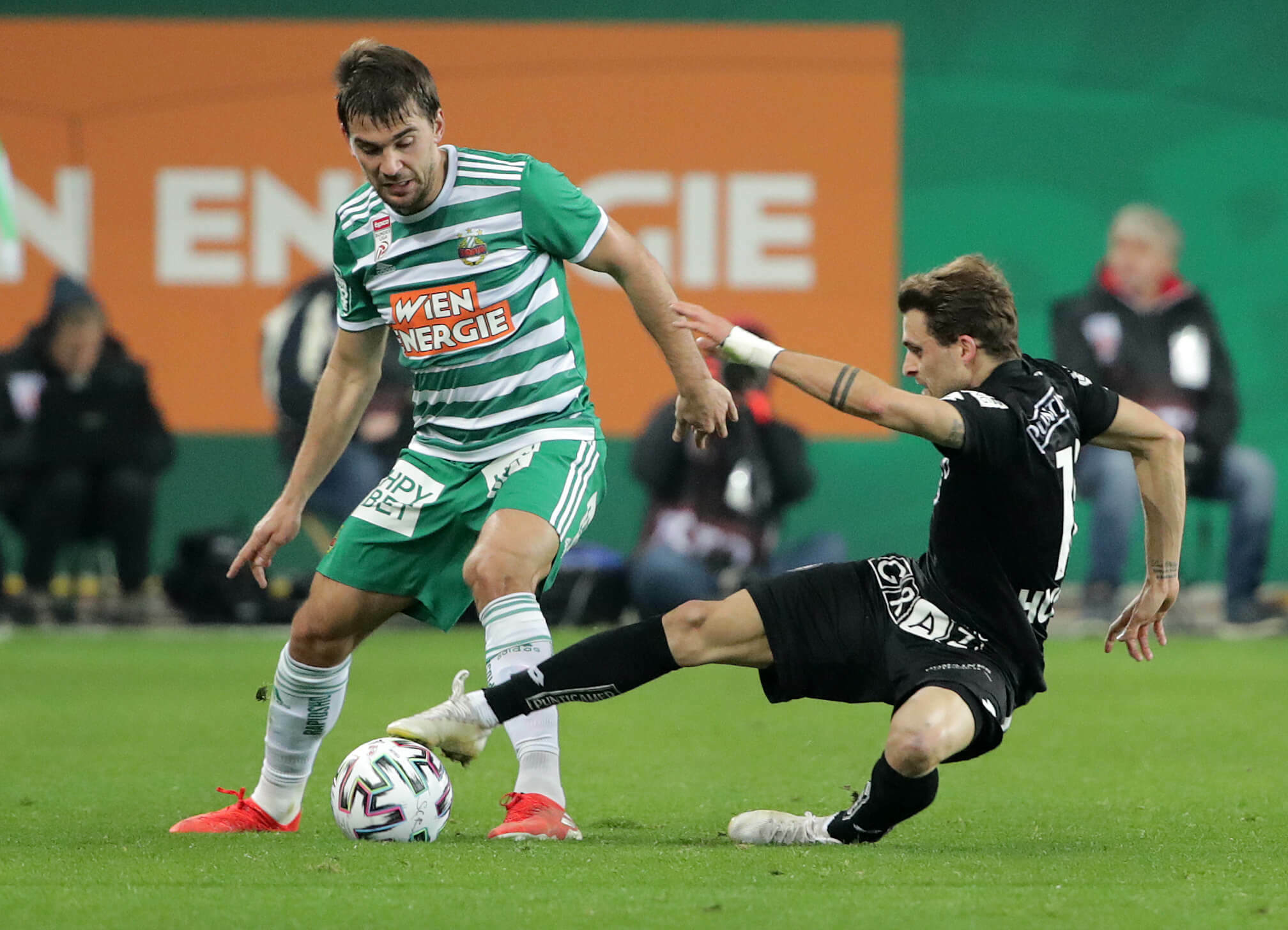 Zwei Fußballspieler mitten in der Aktion; einer in Grün und Weiß dribbelt, der andere in Schwarz rutscht zum Tackling aus.