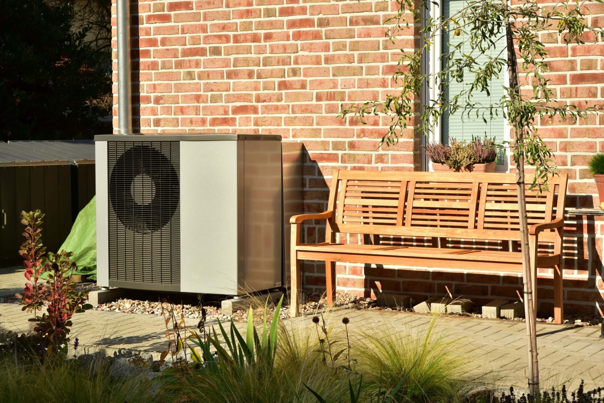 Außenwärmepumpeneinheit neben einer Holzbank auf einer gemauerten Terrasse, umgeben von Pflanzen.