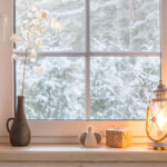 Fenster mit Blick auf einen verschneiten Wald, Vase mit weißen Zweigen, gestricktes Dekor, Geschenkbox und eine brennende Laterne auf der Fensterbank.