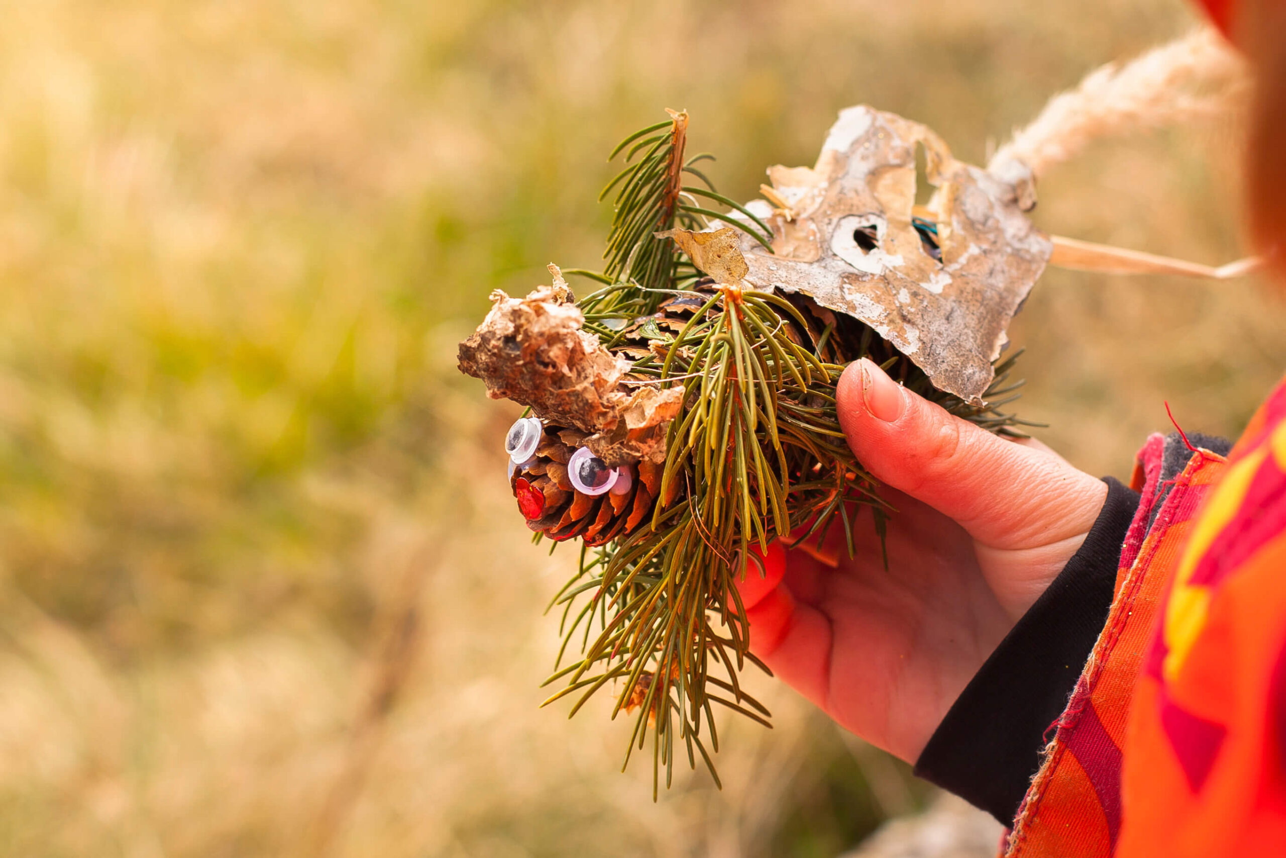 Kind hält ein handgefertigtes Bastelobjekt aus Tannenzapfen, Blättern, Tannennadeln und Wackelaugen.