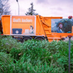 Orangefarbener Müllwagen der MA48 neben einem Stapel Weihnachtsbäume mit einem Sammelstellenschild auf Deutsch.