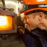 Person mit Schutzhelm und Schutzbrille, die durch ein kleines Fenster in einen Ofen schaut.